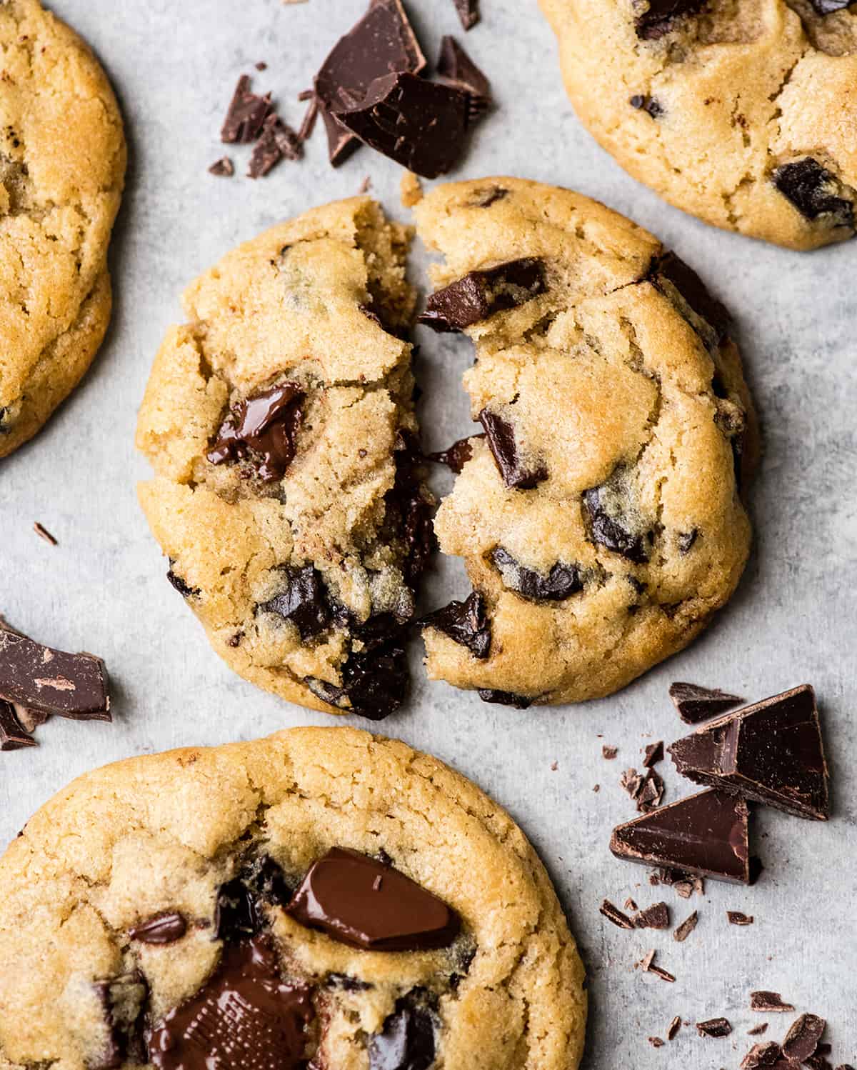 Overhead view of one chocolate chip cookie torn in half with melty chocolate chips surrounded by three other cookies and chopped chocolate