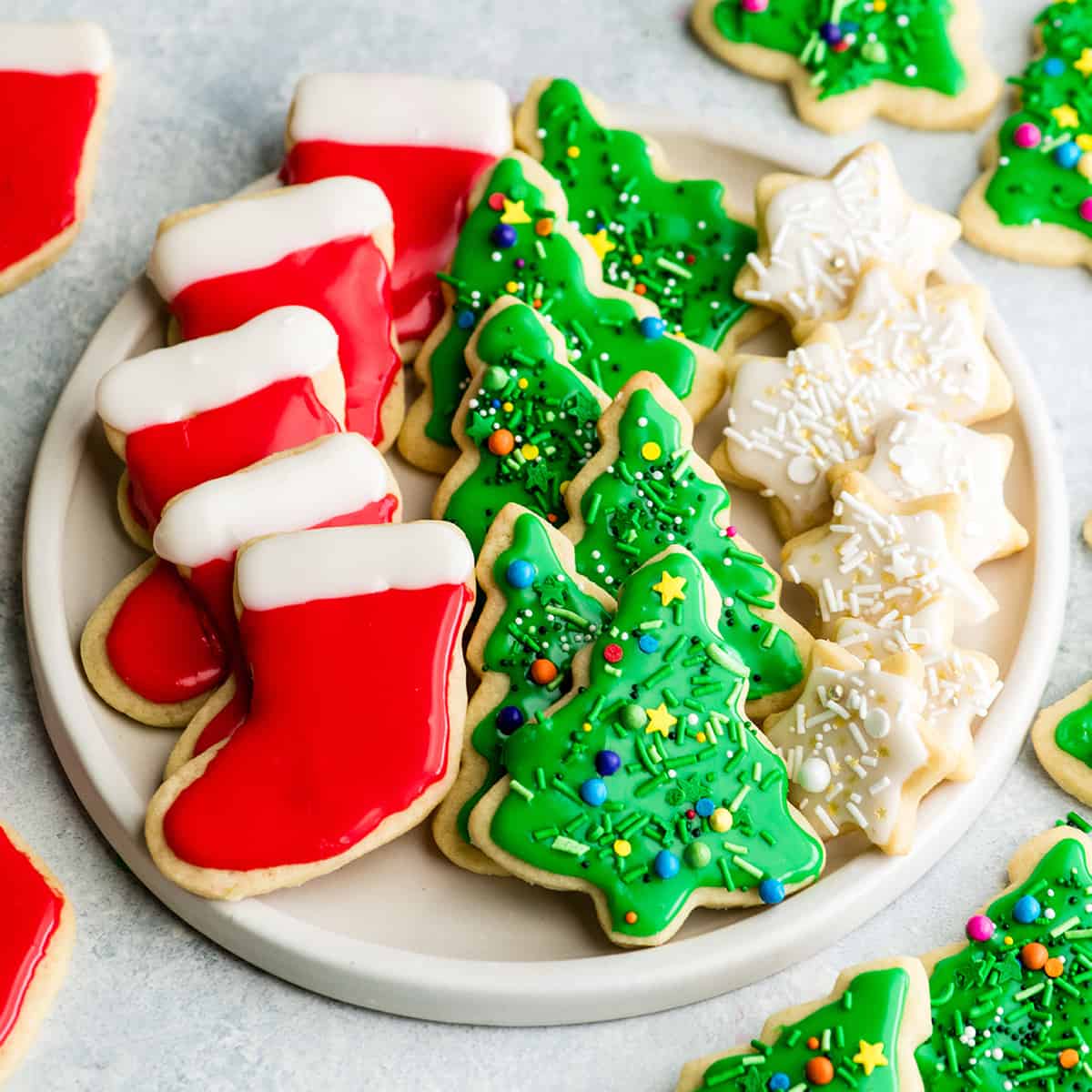 overhead view of a white, round plate with three rows of sugar cookies on it. The left row is five red and white stockings, the middle row is six green Christmas trees with sprinkles and the left row is seven small white stars with sprinkles. 