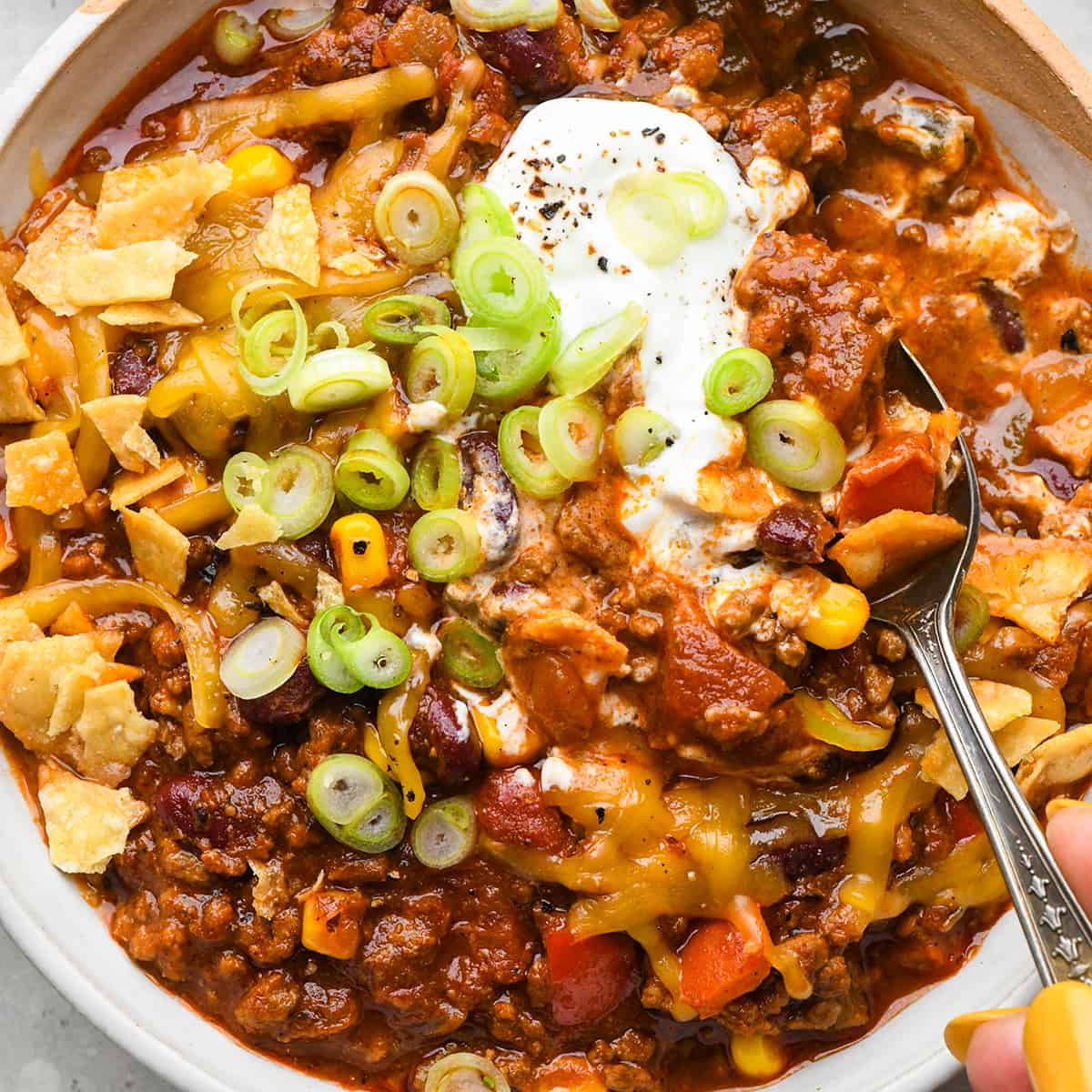 a spoon taking a scoop of beef chili topped with sour cream, green onions, cheese and crushed chips. 