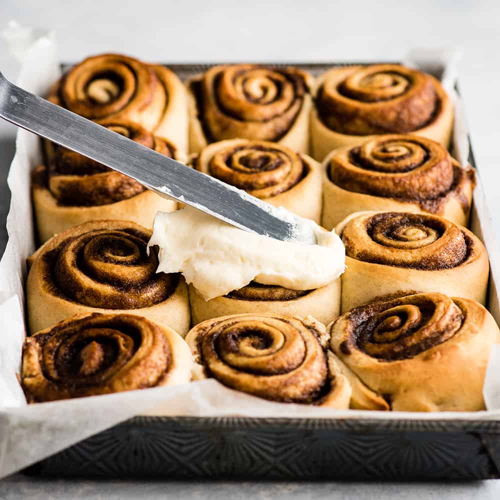 front view of cinnamon rolls being frosted with cream cheese frosting