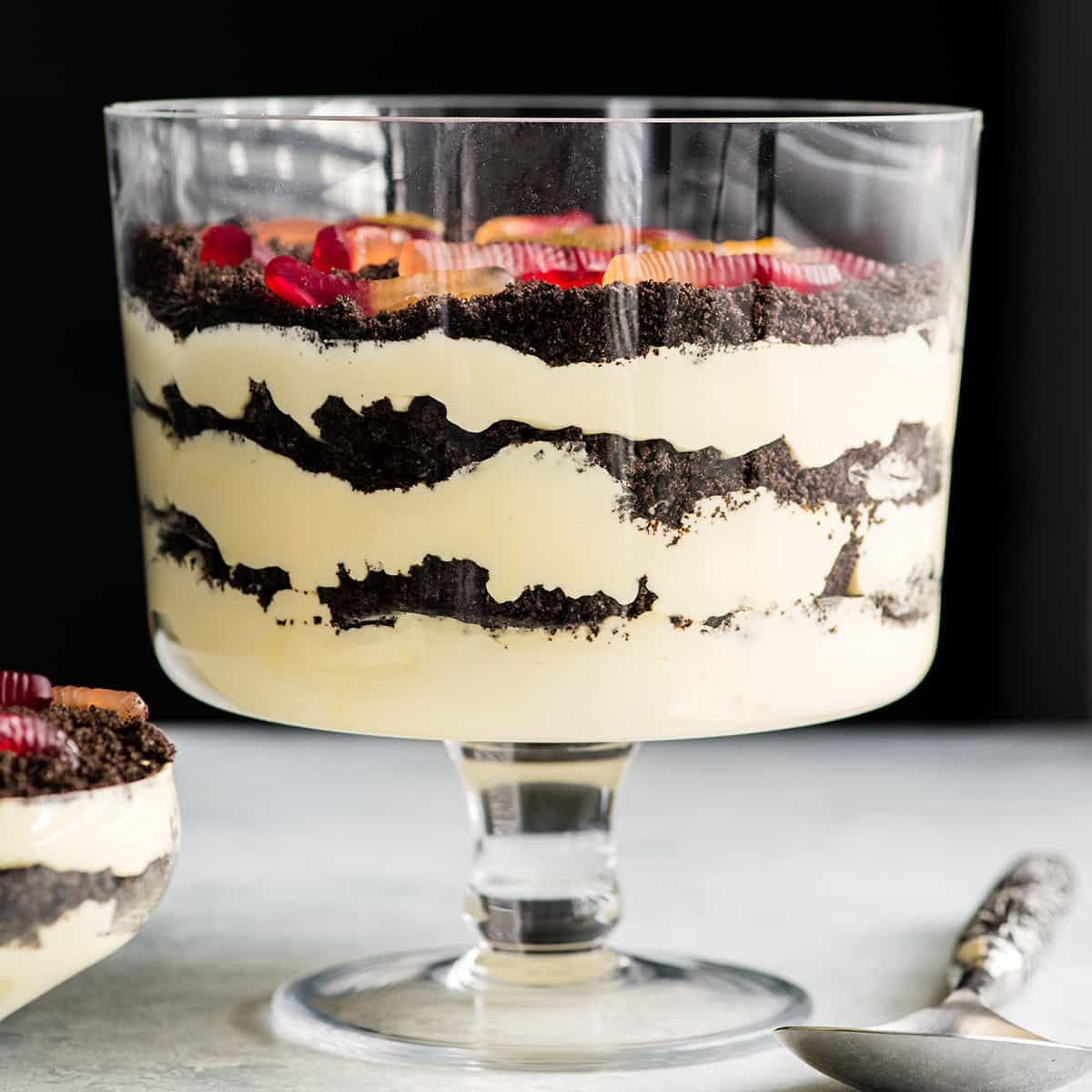 Front view of this Oreo Dirt Cake Recipe in a glass trifle dish with three layers of pudding and three layers of crushed Oreos with gummy worms on top and a spoon and a single-serve dish of Oreo Dirt Cake next to it