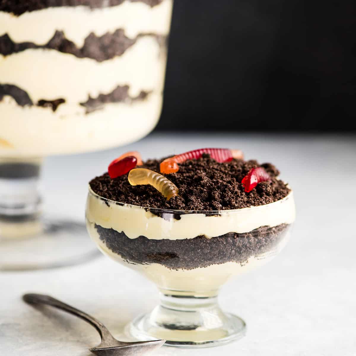 Front view of a small glass dish of Oreo Dirt Cake with two layers of pudding and two layers of crushed oreos with gummy worms on top. A large glass trifle dish of Oreo Dirt Cake is in the background and a silver spoon is in the foreground