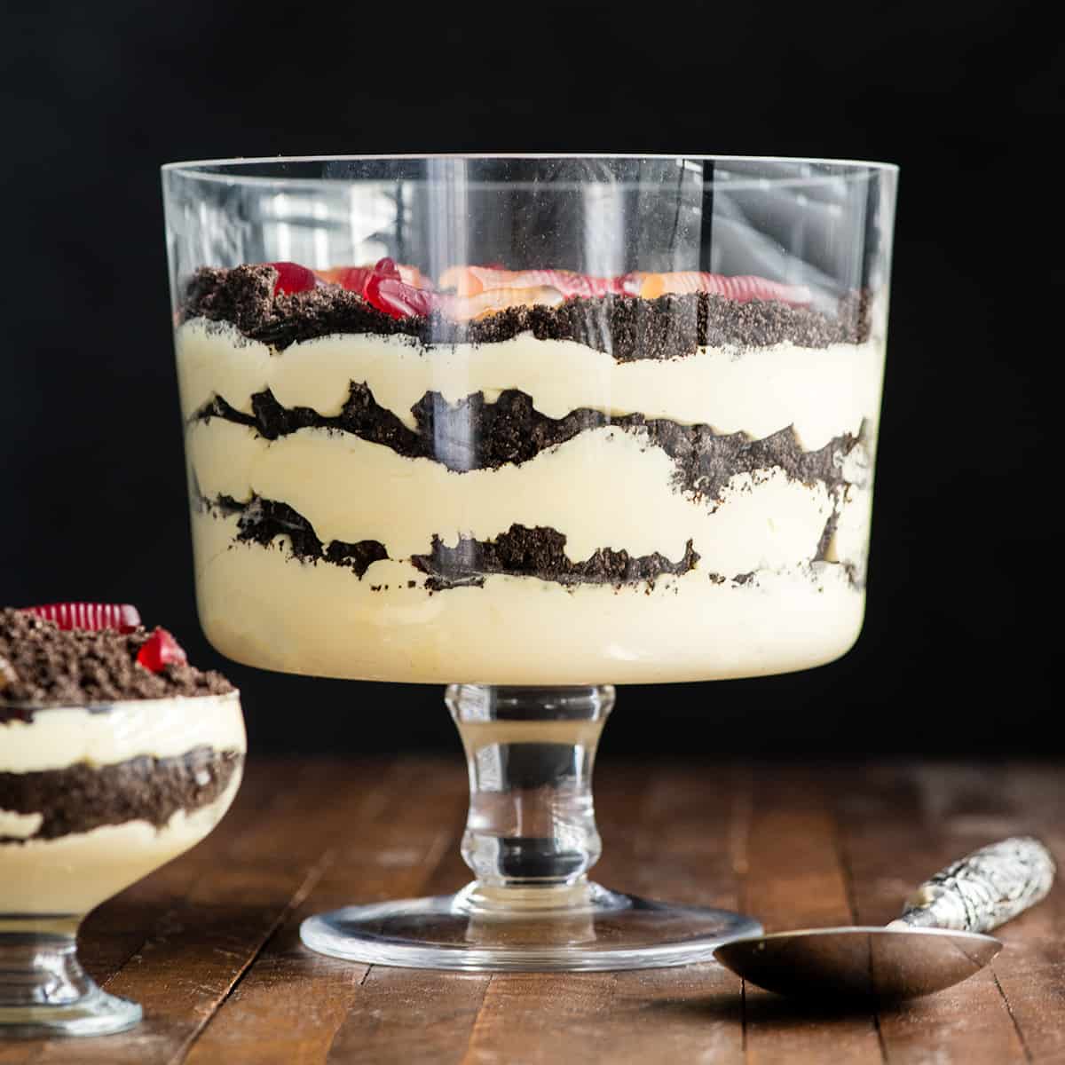 Front view of this Oreo Dirt Cake Recipe in a glass trifle dish with three layers of pudding and three layers of crushed Oreos with gummy worms on top 