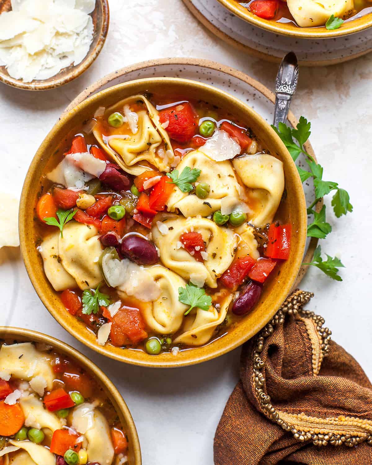 overhead view of a bowl of tortellini soup garnished with parsley and parmesan cheese with two other bowls near it