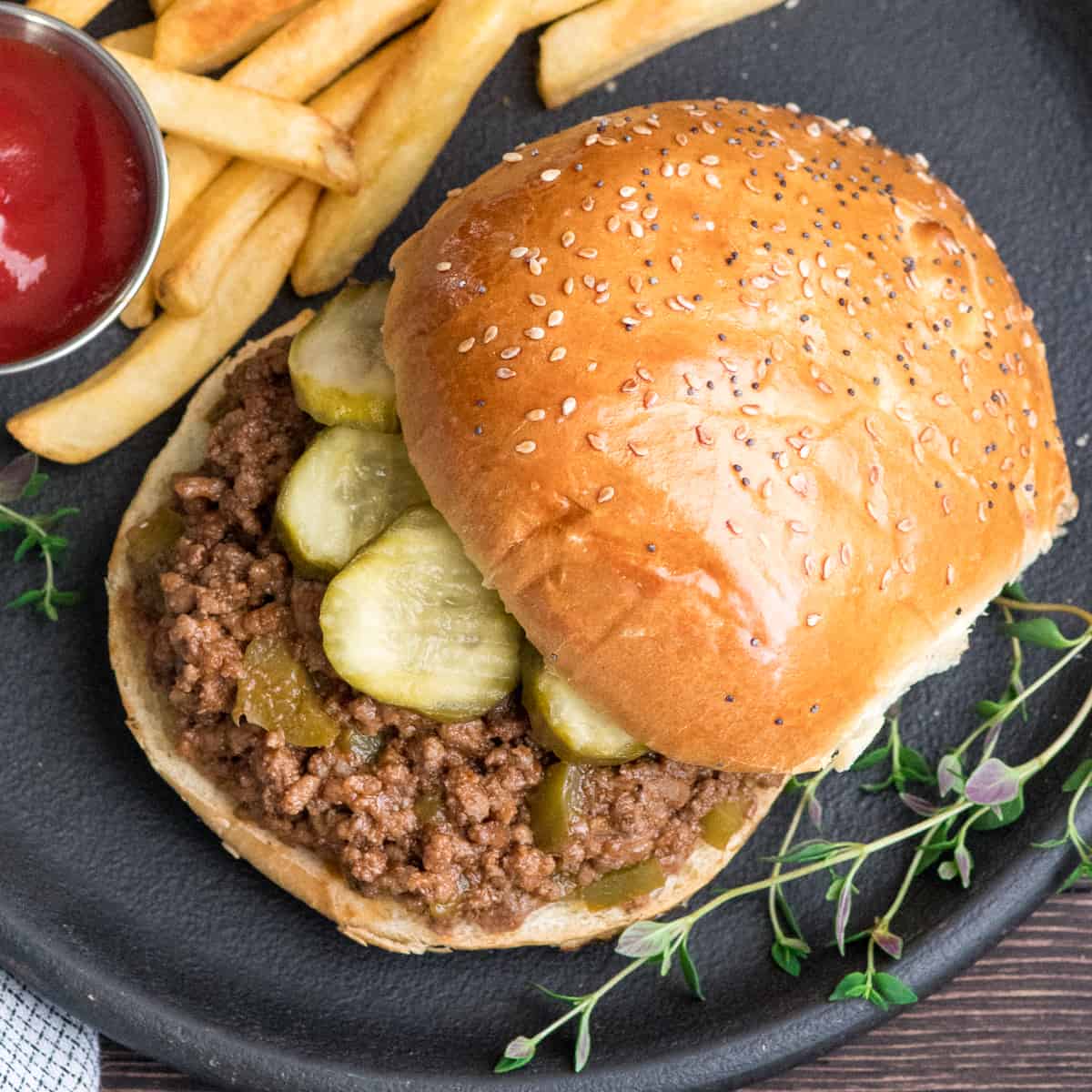 Overhead view of healthy sloppy joes recipe on a bun with pickles on a round black plate with ketchup and french fries and fresh thyme