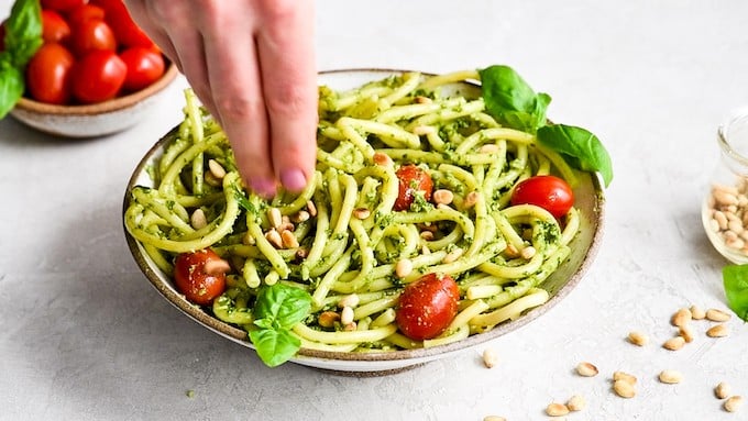 overhead/front view of a hand sprinkling pine nuts over a bowl of Pesto Pasta with tomatoes and fresh basil